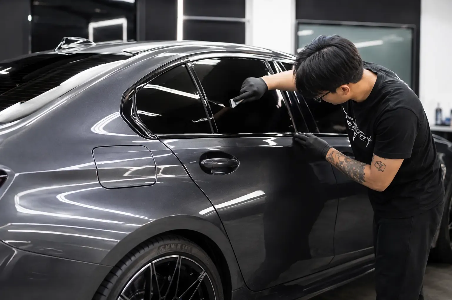 An installer squeegeeing film onto the rear door glass of a gray four-door sedan